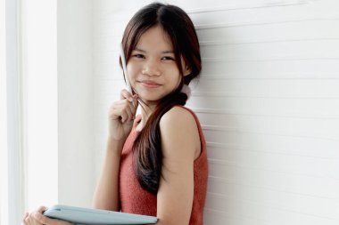 Tablet tutan ve beyaz duvar sınıfında duran mutlu Asyalı kız öğrenci. İlkokul öğrencisi kız çocuğunun portresi okulda öğrenim görüyor. Bayan öğrenci öğrenerek zaman harcıyor. Çocuk ve eğitim