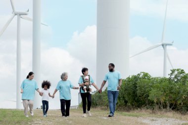 Çeşitlilik ailesi el ele tutuşup rüzgar türbini çiftliğinde çalışıyor. Mutlu ve neşeli insanlar yel değirmeni parkında seyahat ederken birlikte yürürler. Ekoloji doğası yenilenebilir enerji.