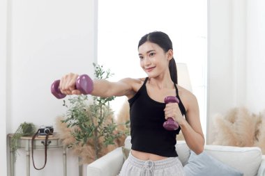 Asyalı güzel kadın evde dambıllarla egzersiz yapıyor. Gülümseyen genç atletik kadın vücudunu eğitmek için dambılını kaldırıyor. Sportif kızlar antrenman yapar. Sağlıklı spor hobi yaşam tarzı.