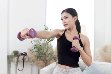 Asyalı güzel kadın evde dambıllarla egzersiz yapıyor. Gülümseyen genç atletik kadın vücudunu eğitmek için dambılını kaldırıyor. Sportif kızlar antrenman yapar. Sağlıklı spor hobi yaşam tarzı.