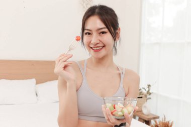 Spor Asyalı kız yatak odasında beyaz yatakta otururken taze meyve ve sebze salatası yiyor. Mutlu güzel kadın evde yemek için çatal ve salata kasesi tutuyor. Sağlıklı yemek ve yaşam tarzı.
