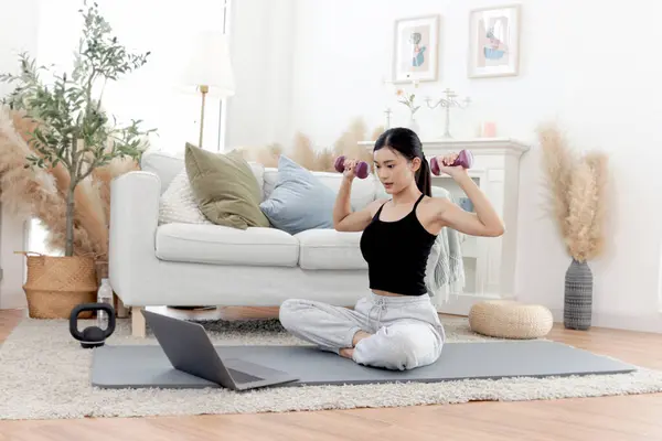 Asian woman lift dumbbells up during watch exercise training tutorial on laptop computer. Happy female work out on yoga mat in home living room. Beautiful sporty girl do workout. Healthy sport hobby.