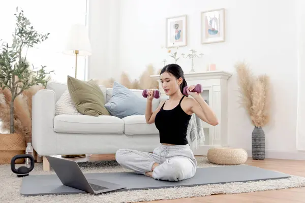 Asian woman lift dumbbells up during watch exercise training tutorial on laptop computer. Happy female work out on yoga mat in home living room. Beautiful sporty girl do workout. Healthy sport hobby.