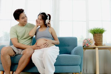 Neşeli Asyalı çift oturma odasındaki kanepede birlikte otururlar. Kulaklıklı güzel hamile kadın, kocasıyla ilgilenirken rahatlamak için müzik dinlemeyi seviyor. Evde mutlu bir aile.