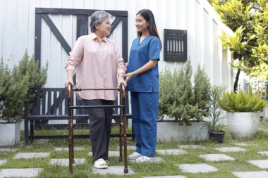 Hemşire, Asyalı kıdemli hastaya dışarıda ortopedi yürüteci ile yürümesi için yardım ediyor. Bahçede fizyoterapist, evde yaşlı hemşire, yaşlı ev bakımı, tıbbi bakım....