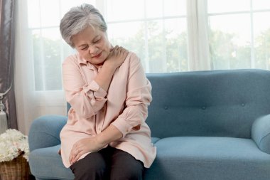 Oturma odasında kanepede otururken omuz ağrısı çeken Asyalı son sınıf öğrencisi bir kadın. Evde omuz ağrısından muzdarip olgunlaşmamış. Mutsuz yaşlı büyükanne omuz ağrısından muzdarip..