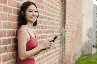 Spor giyimli kısa saçlı spor kulaklıklı kız dışarıda duvara yaslanırken akıllı telefon kullanıyor. Mutlu gülümseyen Asyalı kadın egzersize ara verirken cep telefonu tutuyor.. 