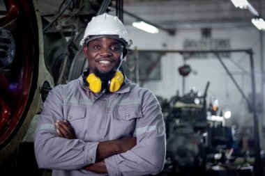 Afrikalı endüstriyel mühendis portresi koruyucu iş kıyafetleri ve güvenli miğfer imalat fabrikasında kolları bağlı duruyor. Metal endüstrisinde çalışan gülümseyen bir erkek işçi..