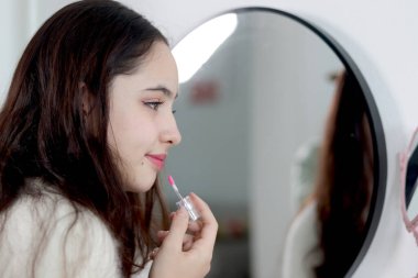 Güzel bir genç kız aynanın önünde ruj tutuyor ve dudaklarına ruj sürüyor. Makyaj yapan güzel bir genç kadın. Her gün güzellik için kozmetik kullanan mutlu genç kız..