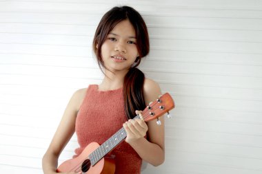 Beyaz odada dururken ukulele tutan Asyalı kız öğrencinin portresi. Müzik aleti olan ilkokul öğrencisi bir kız. Ukulele 'li çocuk.