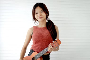 Beyaz odada dururken ukulele tutan Asyalı kız öğrencinin portresi. Müzik aleti olan ilkokul öğrencisi bir kız. Ukulele 'li çocuk.