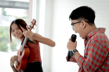 Ukulele tutan Asyalı bir kız ve beyaz odada duran bir çocuk mikrofon tutuyor. İlkokulda beraber müzik çalıp şarkı söylemek yasaktı. Müzik ve enstrümanlı çocuk.