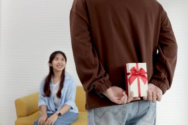 Arkasına hediye kutusu saklayan bir adam. Oturma odasında kutlama yapmak için sevgilisine yıldönümü hediyesi vermeye hazır. Mutlu Asyalı çift sürpriz yapar, aşk ve mutluluk. Sevgililer Günün kutlu olsun..