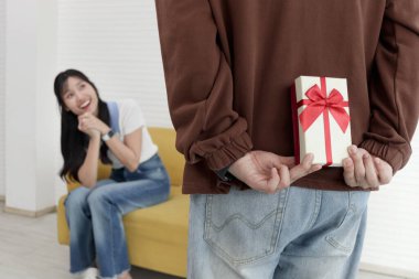 Arkasına hediye kutusu saklayan bir adam. Oturma odasında kutlama yapmak için sevgilisine yıldönümü hediyesi vermeye hazır. Mutlu Asyalı çift sürpriz yapar, aşk ve mutluluk. Sevgililer Günün kutlu olsun..