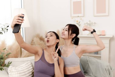 İki sportif Asyalı kız arkadaş oturma odasında kanepede otururken fotoğraf çekmek için cep telefonu kullanıyor. Mutlu güzel bayan arkadaş egzersiz yaptıktan sonra evde selfie çekmek için akıllı telefon kullanıyor..