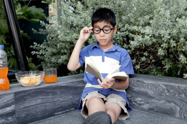 Bahçede gözlüklü Asyalı okul çocuğu kitap okuyor. Öğrenci çocuk parkta kitapla ders öğreniyor. Modern evde eğitim gören odaklanmış bir öğrenci çocuğu. Çocuk her yerde öğrenebilir..