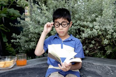 Bahçede gözlüklü Asyalı okul çocuğu kitap okuyor. Öğrenci çocuk parkta kitapla ders öğreniyor. Modern evde eğitim gören odaklanmış bir öğrenci çocuğu. Çocuk her yerde öğrenebilir..