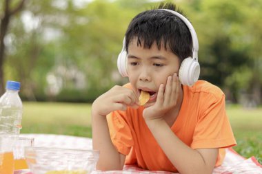 Kulaklıklı mutlu Asyalı okul çocuğu yeşil çimenlerin üzerinde yatarken abur cubur yiyor. Neşeli çocuk güneşli bir günde bahçede vakit geçiriyor. Çocuk yaz parkında piknik yapıyor..