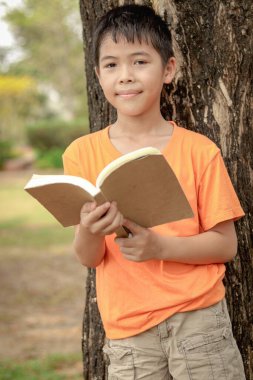 Bahçede kitap okuyan Asyalı okul çocuğunun portresi. Öğrenci çocuk parkta ağacın altında ders çalışıyor. Öğrenci çocuk tek başına öğrenmek için kitap okuyor. Çocuk, eğitim ve doğa..