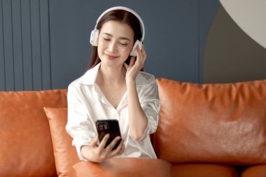 Oturma odasında kanepede otururken müzik dinlemek için cep telefonu kullanan orta yaşlı mutlu bir kadın. Güzel, rahatlatıcı yetişkin Asyalı kadın özgürlüğünü evde müzik dinleyerek geçiriyor..