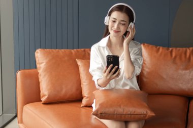 Oturma odasında kanepede otururken müzik dinlemek için cep telefonu kullanan orta yaşlı mutlu bir kadın. Güzel, rahatlatıcı yetişkin Asyalı kadın özgürlüğünü evde müzik dinleyerek geçiriyor..