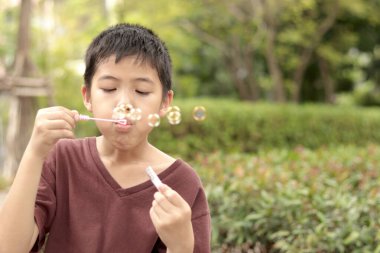 Güneşli bir günde parkta baloncuk çıkaran mutlu Asyalı çocuk. Küçük çocuk yeşil bahçede baloncuk üflüyor. Neşeli çocuk dışarıda doğayla oynamaktan hoşlanıyor. Çocukluk açık hava konsepti.