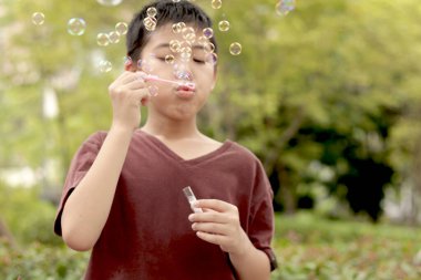 Güneşli bir günde parkta baloncuk çıkaran mutlu Asyalı çocuk. Küçük çocuk yeşil bahçede baloncuk üflüyor. Neşeli çocuk dışarıda doğayla oynamaktan hoşlanıyor. Çocukluk açık hava konsepti.