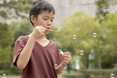 Güneşli bir günde parkta baloncuk çıkaran mutlu Asyalı çocuk. Küçük çocuk yeşil bahçede baloncuk üflüyor. Neşeli çocuk dışarıda doğayla oynamaktan hoşlanıyor. Çocukluk açık hava konsepti.