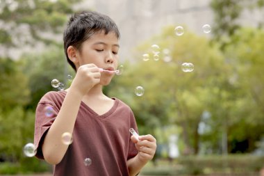 Güneşli bir günde parkta baloncuk çıkaran mutlu Asyalı çocuk. Küçük çocuk yeşil bahçede baloncuk üflüyor. Neşeli çocuk dışarıda doğayla oynamaktan hoşlanıyor. Çocukluk açık hava konsepti.