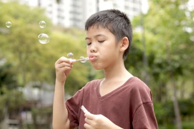 Güneşli bir günde parkta baloncuk çıkaran mutlu Asyalı çocuk. Küçük çocuk yeşil bahçede baloncuk üflüyor. Neşeli çocuk dışarıda doğayla oynamaktan hoşlanıyor. Çocukluk açık hava konsepti.