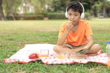 Kulaklıklı mutlu Asyalı okul çocuğu yeşil çimlerin üzerinde otururken abur cubur yiyor. Neşeli çocuk güneşli bir günde bahçede vakit geçiriyor. Çocuk yaz parkında piknik yapıyor..