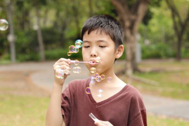 Güneşli bir günde parkta baloncuk çıkaran mutlu Asyalı çocuk. Küçük çocuk yeşil bahçede baloncuk üflüyor. Neşeli çocuk dışarıda doğayla oynamaktan hoşlanıyor. Çocukluk açık hava konsepti.
