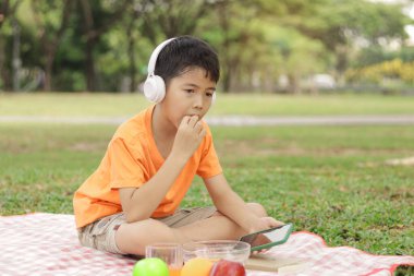 Kulaklıklı mutlu Asyalı okul çocuğu yeşil çimlerin üzerinde otururken abur cubur yiyor. Neşeli çocuk güneşli bir günde bahçede vakit geçiriyor. Çocuk yaz parkında piknik yapıyor..