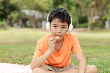 Kulaklıklı mutlu Asyalı okul çocuğu yeşil çimlerin üzerinde otururken abur cubur yiyor. Neşeli çocuk güneşli bir günde bahçede vakit geçiriyor. Çocuk yaz parkında piknik yapıyor..
