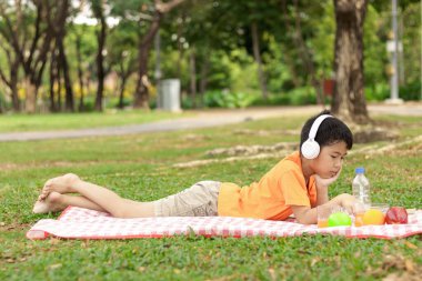 Kulaklıklı mutlu Asyalı okul çocuğu yeşil çimenlerin üzerinde yatarken abur cubur yiyor. Neşeli çocuk güneşli bir günde bahçede vakit geçiriyor. Çocuk yaz parkında piknik yapıyor..