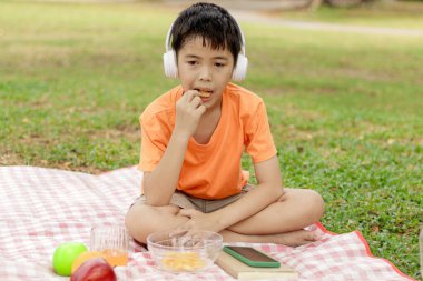 Kulaklıklı mutlu Asyalı okul çocuğu yeşil çimlerin üzerinde otururken abur cubur yiyor. Neşeli çocuk güneşli bir günde bahçede vakit geçiriyor. Çocuk yaz parkında piknik yapıyor..