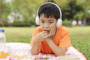 Kulaklıklı mutlu Asyalı okul çocuğu yeşil çimenlerin üzerinde yatarken abur cubur yiyor. Neşeli çocuk güneşli bir günde bahçede vakit geçiriyor. Çocuk yaz parkında piknik yapıyor..