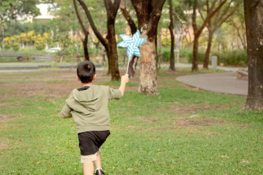 Güneşli bir günde parkta rüzgar türbiniyle oynayan mutlu Asyalı çocuk. Doğada rüzgar türbini olan neşeli bir çocuk. Neşeli küçük çocuk bahçede vakit geçiriyor. Çocukluk açık hava konsepti.