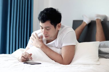 Asyalı bir adam sabah yatak odasında uyandıktan sonra yatakta akıllı telefon kullanıyor. Rahat bir yatakta uzanmış cep telefonunu kontrol eden bir adam. Uykucu adam evde akıllı telefonu tutuyor..