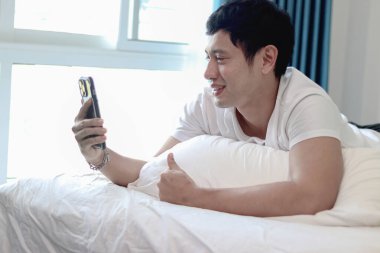 Asyalı bir adam sabah uyandıktan sonra yatak odasında akıllı telefon kullanıyor. Rahat bir yatakta uzanmış cep telefonunu kontrol eden bir adam. Uykucu adam evde akıllı telefonu tutuyor..