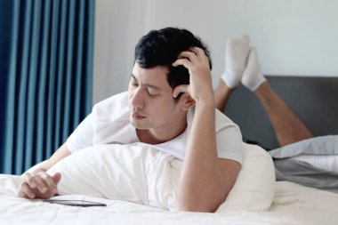 Asyalı bir adam sabah yatak odasında uyandıktan sonra yatakta akıllı telefon kullanıyor. Rahat bir yatakta uzanmış cep telefonunu kontrol eden bir adam. Uykucu adam evde akıllı telefonu tutuyor..