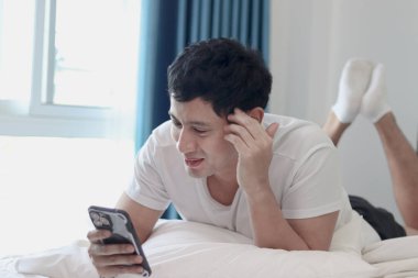 Asyalı bir adam sabah yatak odasında uyandıktan sonra yatakta akıllı telefon kullanıyor. Rahat bir yatakta uzanmış cep telefonunu kontrol eden bir adam. Uykucu adam evde akıllı telefonu tutuyor..