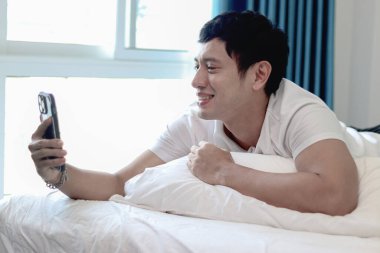 Asyalı bir adam sabah yatak odasında uyandıktan sonra yatakta akıllı telefon kullanıyor. Rahat bir yatakta uzanmış cep telefonunu kontrol eden bir adam. Uykucu adam evde akıllı telefonu tutuyor..