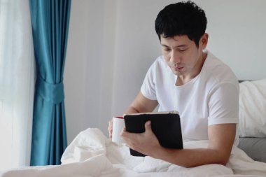 Asyalı adam sabah yatak odasında uyandıktan sonra yatakta kahve içerken tablet kullanıyor. Adam rahat bir yatakta yatarken tabletinin üzerinde çalışıyor. Uykulu adam evde yatakta internette sörf yapıyor..