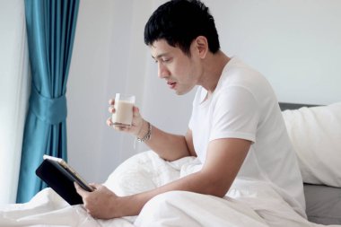 Asyalı bir adam sabah yatak odasında uyandıktan sonra yatakta badem sütü içerken tablet kullanıyor. Adam rahat bir yatakta yatarken tabletinin üzerinde çalışıyor. Uykulu adam yatakta internette sörf yapıyor..