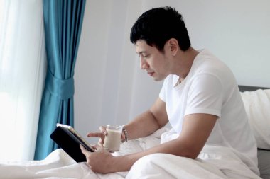 Asyalı bir adam sabah yatak odasında uyandıktan sonra yatakta badem sütü içerken tablet kullanıyor. Adam rahat bir yatakta yatarken tabletinin üzerinde çalışıyor. Uykulu adam yatakta internette sörf yapıyor..