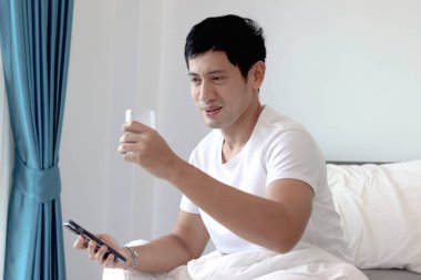 Asyalı bir adam sabah yatak odasında uyandıktan sonra yatakta badem sütü içerken akıllı telefon kullanıyor. Evde rahat bir yatakta uzanırken içeceğe ve cep telefonuna bakan bir adam..