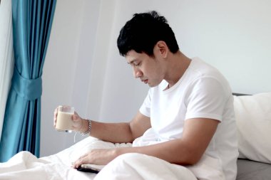 Asyalı bir adam sabah yatak odasında uyandıktan sonra yatakta badem sütü içerken tablet kullanıyor. Adam rahat bir yatakta yatarken tabletinin üzerinde çalışıyor. Uykulu adam yatakta internette sörf yapıyor..