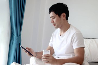 Asyalı bir adam sabah yatak odasında uyandıktan sonra yatakta badem sütü içerken akıllı telefon kullanıyor. Adam rahat bir yatakta uzanırken cep telefonunu kontrol ediyor. Uykulu adam internette sörf yapıyor.