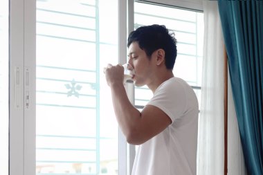 Asyalı bir adam sabah kalktıktan sonra yatak odasının penceresinde badem sütü içiyor. Yatak odasında süt bardağı tutan adam. Uykulu adam sağlıklı içeceğini evde içer..